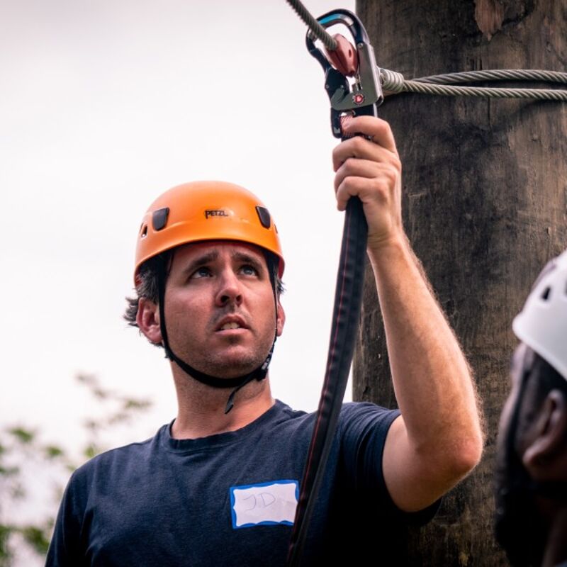 The image shows a man wearing an orange helmet, looking up with a focused expression. He is holding a black strap connected to a metal carabiner, likely part of a zip-lining or climbing setup. The background is slightly blurred, suggesting an outdoor environment. Another person wearing a white helmet is partially visible on the right side of the image.